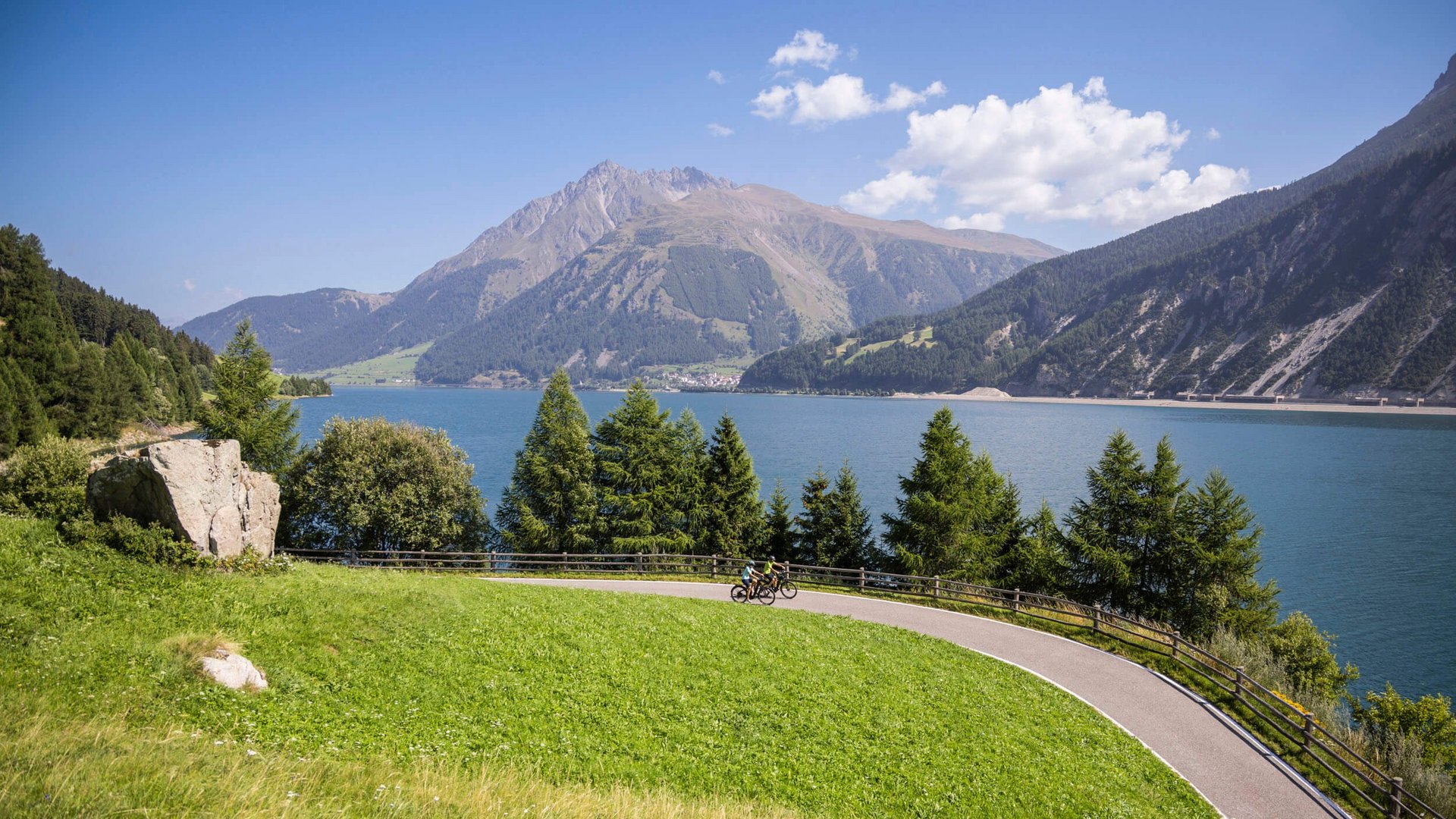 Radfahrer auf einem geschwungenen Weg am See, umgeben von grünen Hängen und Bergen (© Velontour - Heiko Mandl)