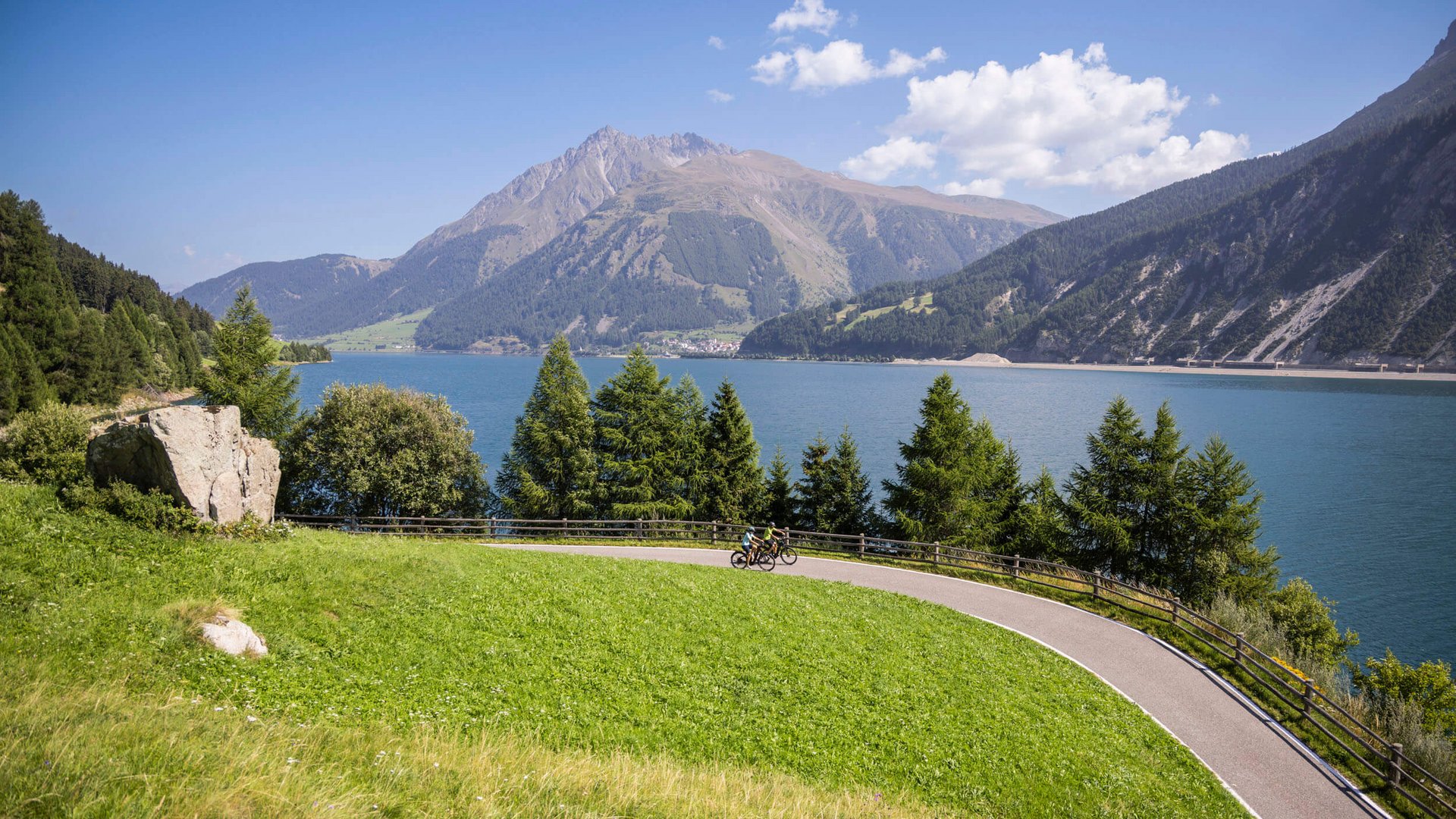 Radfahrer auf einem geschwungenen Weg am See, umgeben von grünen Hängen und Bergen (© Velontour - Heiko Mandl)